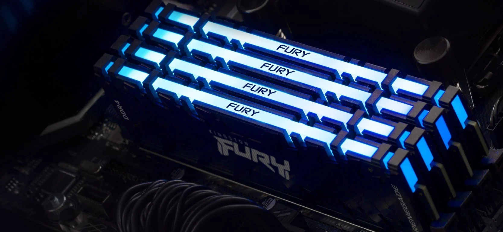 Quatro pentes de memória RAM Kingston FURY Renegade RGB instalados em uma placa-mãe, com iluminação LED azul brilhante em um setup gamer.