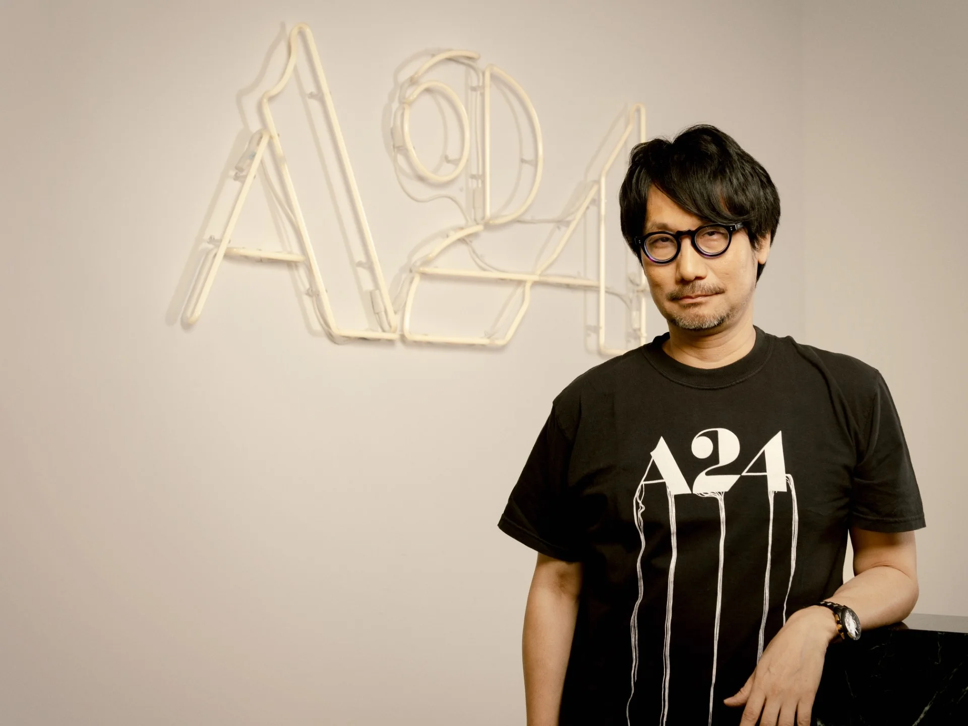 Hideo Kojima ao lado do logotipo da A24 em neon, na foto oficial que anuncia a adaptação de Death Stranding para o cinema